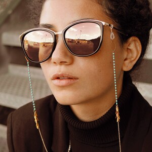 May include: A woman wearing a black turtleneck sweater and sunglasses with a turquoise beaded chain. The sunglasses have a rose gold frame and mirrored lenses.