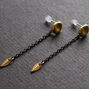 May include: A pair of gold earrings with black chain links and a leaf-shaped charm at the bottom. The earrings have a round, disc-shaped top.