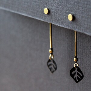 May include: A pair of gold earrings with black leaf-shaped charms hanging from them. The earrings are displayed on a gray surface.