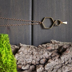 May include: A gold necklace with a geometric pendant featuring three connected hexagonal shapes. The necklace is displayed on a wooden surface with green moss.