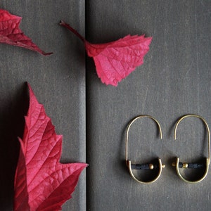 May include: A pair of gold hoop earrings with black beads and gold accents. The earrings are on a dark brown wooden surface with red leaves.