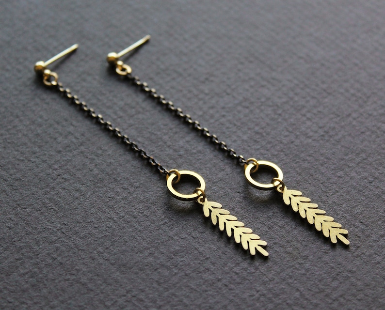 May include: A pair of gold dangle earrings with a black chain and a leaf design. The earrings have a small circle at the top and a long, thin chain that ends in a leaf-shaped pendant.