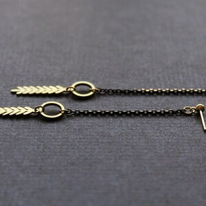 May include: A pair of gold earrings with a long chain and a small arrow-shaped pendant. The chain is black and the pendant is gold.