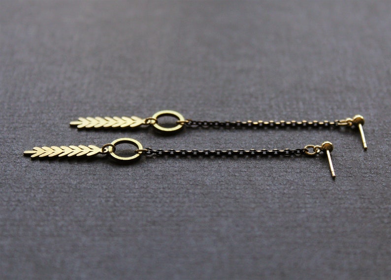 May include: A pair of gold earrings with a long chain and a small arrow-shaped pendant. The chain is black and the pendant is gold.