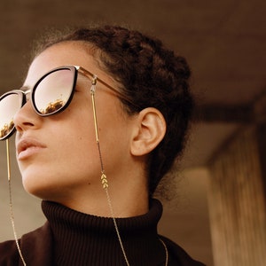 May include: A person wearing black framed sunglasses with a gold chain attached to the temples. The chain has small gold arrow-shaped charms. The person is wearing a black turtleneck sweater.