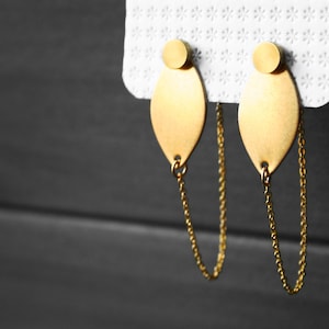May include: A pair of gold-toned earrings with a leaf-shaped pendant and a chain that hangs down from the bottom. The earrings are on a white card with a pattern of small stars.