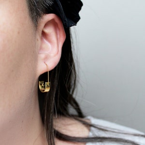 May include: Gold hoop earrings with a unique, slightly curved design. The earrings are worn by a person with short, dark hair.