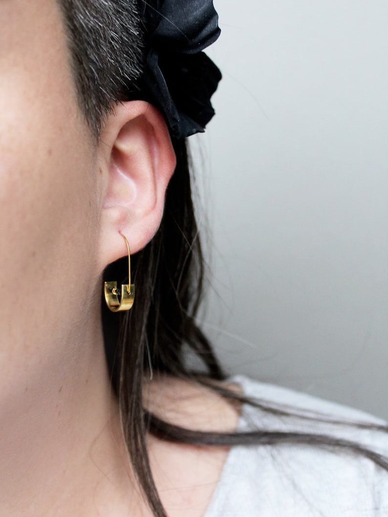 May include: Gold hoop earrings with a unique, slightly curved design. The earrings are worn by a person with short, dark hair.