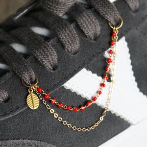 May include: Close-up of a grey suede trainer with white detailing. A gold-coloured shoe charm with a leaf pendant and red beaded chain hangs from the laces, adding a touch of elegance to the casual footwear.
