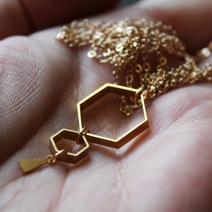 May include: A gold necklace with a geometric pendant featuring two connected hexagonal shapes. The pendant is suspended from a delicate gold chain.