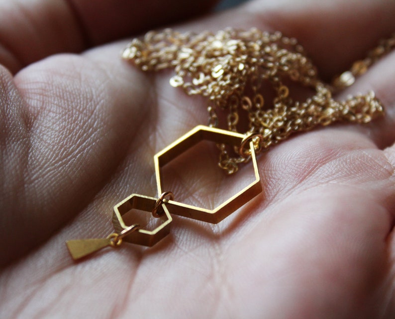 May include: A gold necklace with a geometric pendant featuring two connected hexagonal shapes. The pendant is suspended from a delicate gold chain.