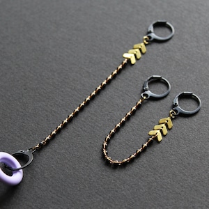 May include: Two pairs of black hoop earrings with gold arrow charms hanging from them. The earrings are on a gray background.