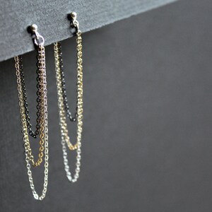 May include: Three pairs of chain earrings in gold, silver, and black hanging from a gray surface.