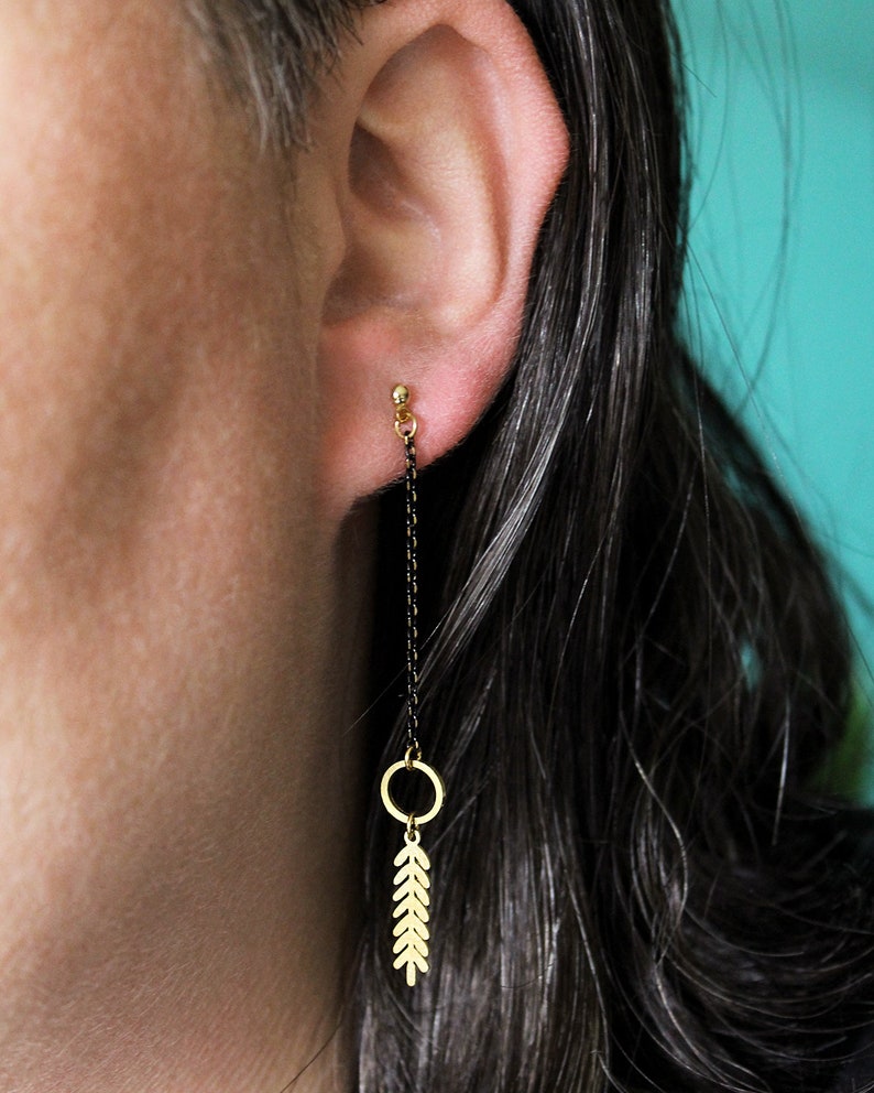 May include: Gold leaf earrings with a black chain and a small gold circle. The earrings are hanging from the earlobe.