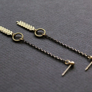 May include: A pair of gold earrings with a leaf design and a small circle dangle. The earrings are attached to a black chain with a small ball at the end.