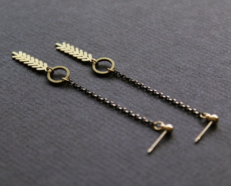 May include: A pair of gold earrings with a leaf design and a small circle dangle. The earrings are attached to a black chain with a small ball at the end.