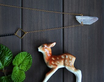 May include: A gold chain necklace with a clear quartz crystal pendant and a square charm. The necklace is displayed on a dark wood surface with a small ceramic deer figurine.