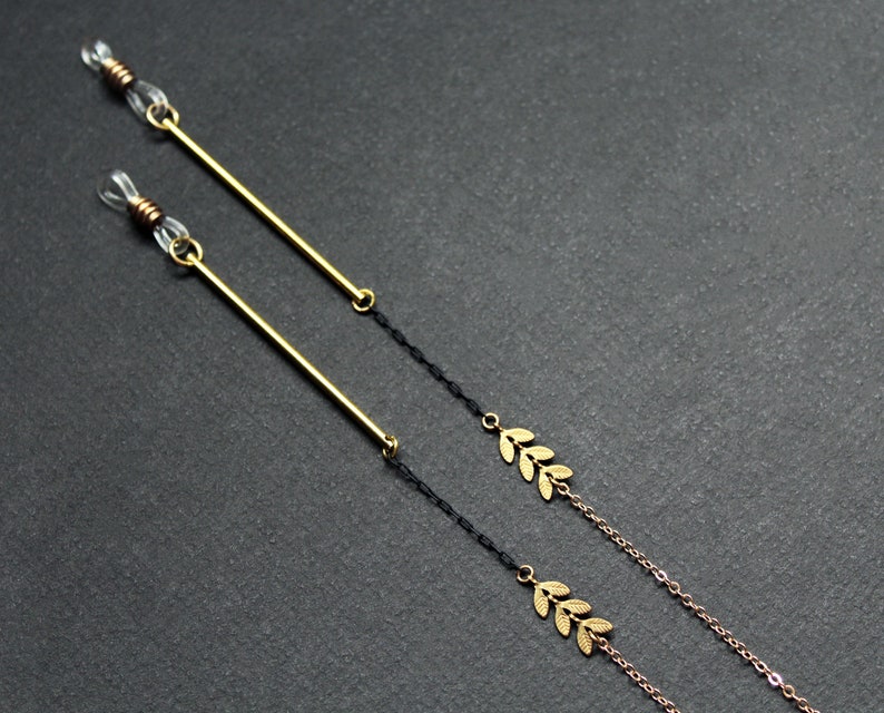 May include: Two gold eyeglass chains with black cord and gold leaf accents. The chains are on a gray background.