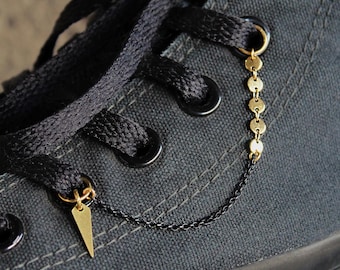 May include: Close-up of a black canvas shoe with black laces. Gold-colored shoe charms are attached to the laces, including a small triangle and a chain of connected circles. The charms add a decorative element to the shoe.