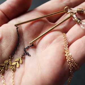 May include: A gold necklace with a geometric pendant and a chain of small gold leaves. The pendant is made of two thin gold bars connected at the top and bottom. The necklace is on a hand.