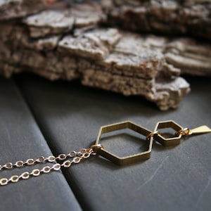 May include: A gold necklace with a geometric pendant made of two connected hexagonal shapes. The pendant has a small, pointed detail at the top.