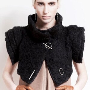 May include: A black cropped jacket with a high collar and decorative silver hardware. The jacket has a textured, fuzzy surface and is worn over a light brown dress.