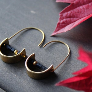 May include: A pair of gold hoop earrings with black beads. The earrings are on a gray surface with a red leaf in the background.