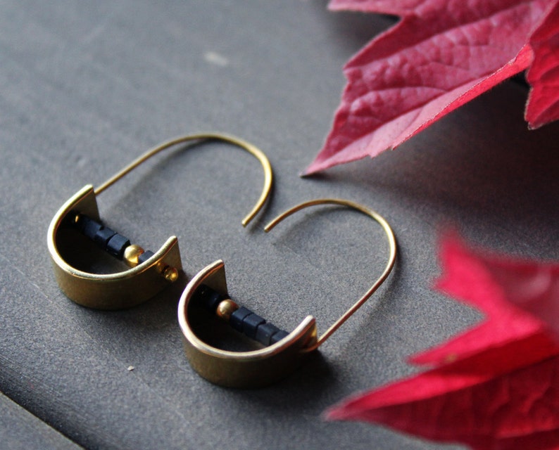 May include: A pair of gold hoop earrings with black beads. The earrings are on a gray surface with a red leaf in the background.