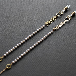 May include: A pair of gold-tone eyeglass chains with white pearl beads and leaf-shaped charms. The chains are attached to clear silicone loops.