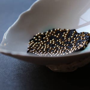 May include: A white ceramic bowl filled with black and gold beads. The beads are arranged in a pattern, creating a visually appealing design.
