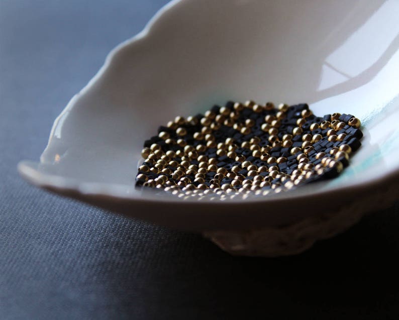 May include: A white ceramic bowl filled with black and gold beads. The beads are arranged in a pattern, creating a visually appealing design.