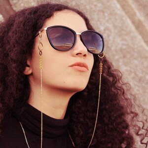 K&ouml;nnte beinhalten: Eine Frau mit lockigem braunen Haar tr&auml;gt eine schwarze Sonnenbrille mit einer goldenen Kette, die an den B&uuml;geln befestigt ist. Die Kette hat goldene Perlen und h&auml;ngt bis zu ihrer Brust.