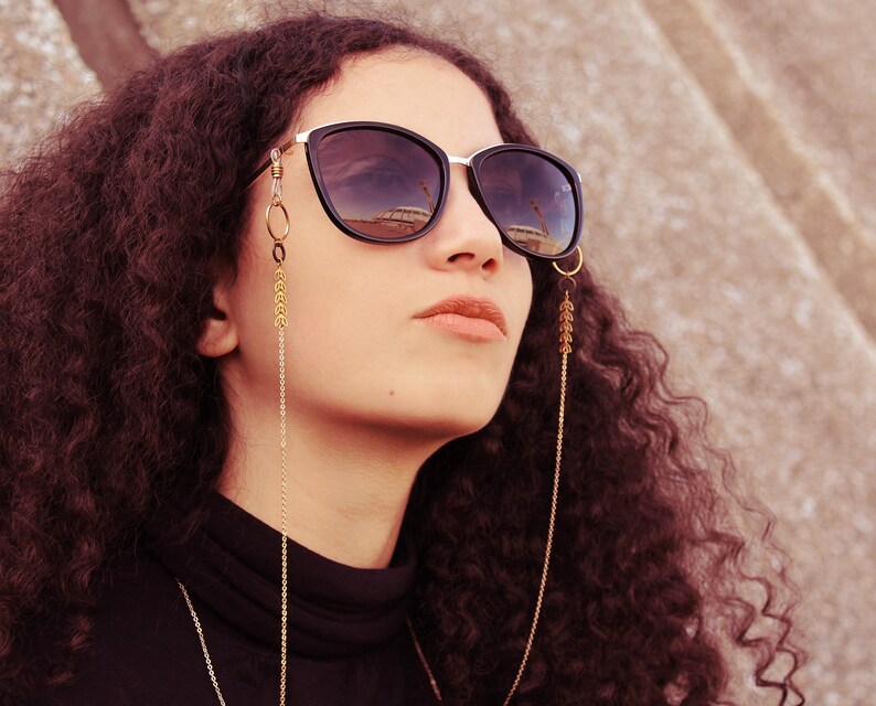 K&ouml;nnte beinhalten: Eine Frau mit lockigem braunen Haar tr&auml;gt eine schwarze Sonnenbrille mit einer goldenen Kette, die an den B&uuml;geln befestigt ist. Die Kette hat goldene Perlen und h&auml;ngt bis zu ihrer Brust.