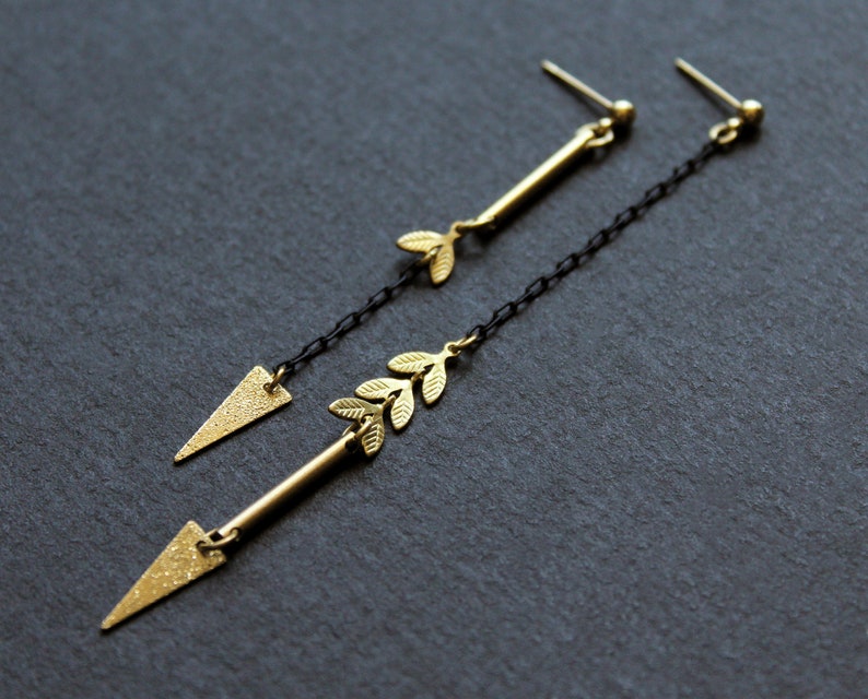 May include: A pair of gold earrings with a delicate leaf design and a triangle shape at the bottom. The earrings are suspended from a black chain.