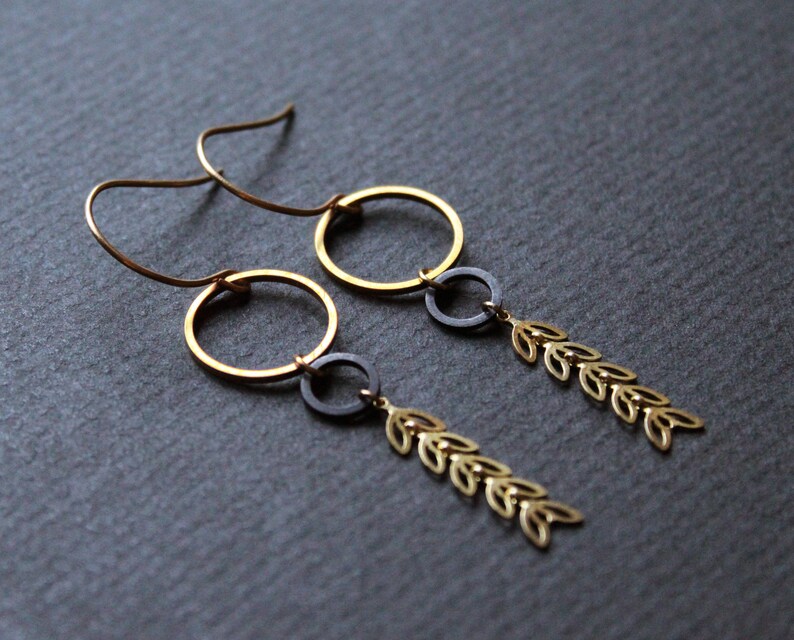 K&ouml;nnte beinhalten: Ein Paar goldene und schwarze Ohrringe mit einem Kreisdesign und einer herabh&auml;ngenden Kette aus kleinen Bl&auml;ttern.