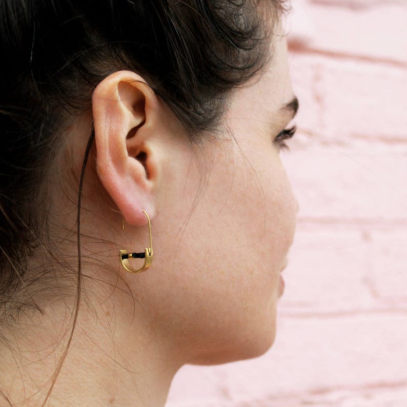 May include: A person wearing a gold hoop earring with a black accent. The earring is a simple design with a curved shape.