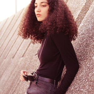 May include: A woman with curly brown hair wearing a black mock neck shirt and blue jeans. She is holding a pair of sunglasses attached to a gold chain.