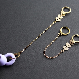 May include: Two pairs of gold earrings connected by a chain. One pair has a purple silicone earplug attached to the chain. The earrings feature small heart-shaped charms.