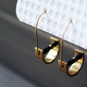 May include: A pair of gold hoop earrings with black cord accents. The hoops are open and have a simple, minimalist design.