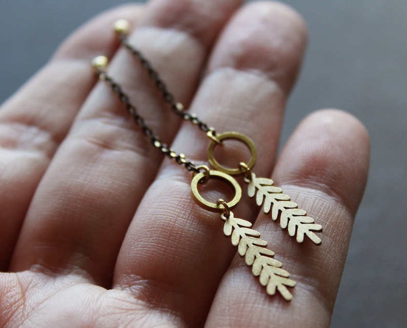 May include: A pair of gold-toned earrings with a delicate chain and a leaf-shaped pendant. The earrings are hanging from a hand.