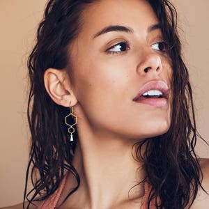 May include: A woman with wet, dark hair wearing a peach-colored halter top and gold geometric earrings. The earrings feature three hexagonal shapes and a small tassel.