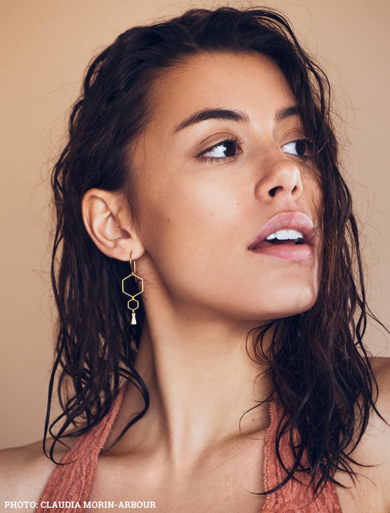 May include: A woman with wet, dark hair wearing a peach-colored halter top and gold geometric earrings. The earrings feature three hexagonal shapes and a small tassel.