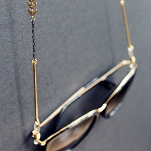 May include: A pair of black sunglasses with gold accents hanging from a gold chain with a leaf design. The chain is attached to the sunglasses with gold hooks.