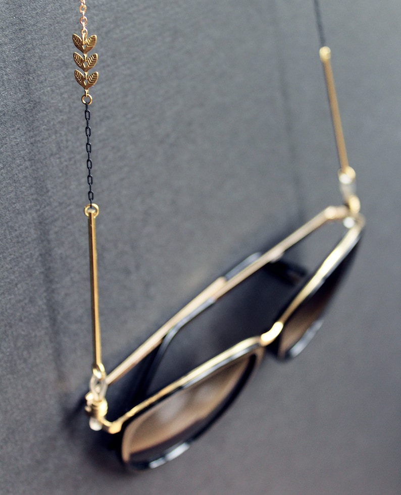 May include: A pair of black sunglasses with gold accents hanging from a gold chain with a leaf design. The chain is attached to the sunglasses with gold hooks.