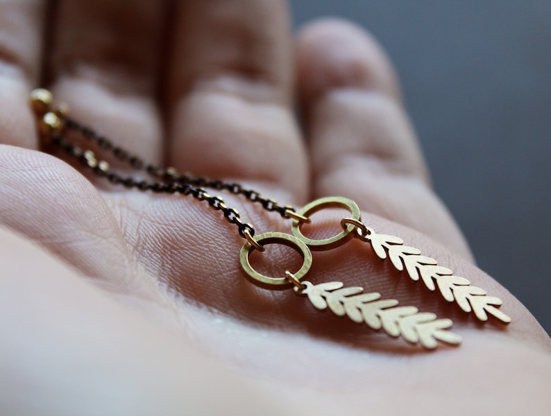May include: A pair of gold earrings with delicate leaf-shaped charms hanging from small hoops. The earrings are on a hand, with the chain visible.
