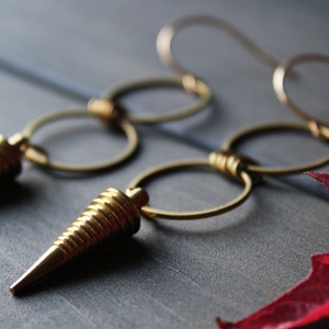 May include: Gold-toned earrings featuring a series of linked circles and cone-shaped pendants. The earrings have a textured, ridged design on the cone-shaped elements. The background is a muted gray with a hint of red from a leaf.