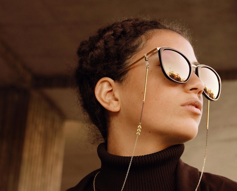May include: A person wearing black sunglasses with a gold chain attached to the temples. The chain has small leaf-shaped charms.