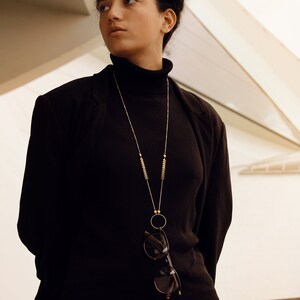 May include: A woman wearing a black turtleneck sweater and a black blazer. She is wearing a gold chain necklace with a small circle pendant and a pair of glasses hanging from the chain.