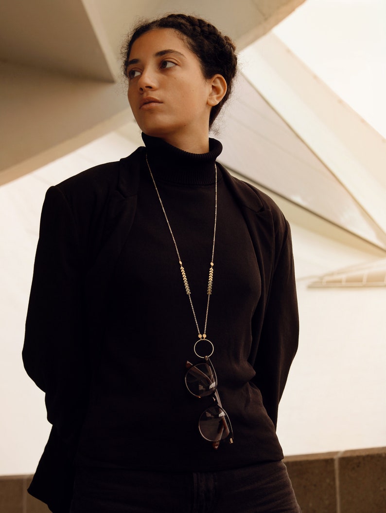 May include: A woman wearing a black turtleneck sweater and a black blazer. She is wearing a gold chain necklace with a small circle pendant and a pair of glasses hanging from the chain.