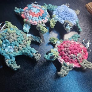 May include: Four crocheted turtle keychains in different colors: green, blue, pink, and multi-colored. The turtles are all facing the same direction with their heads up.
